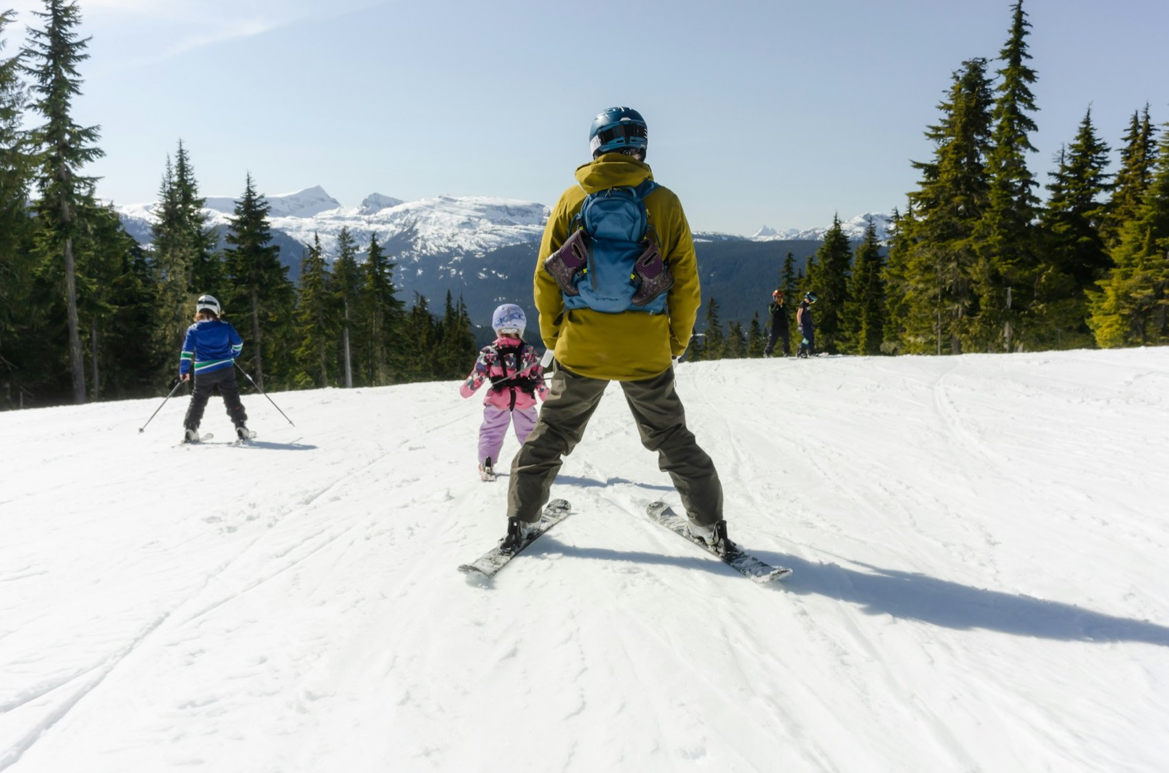 A parent and two children skiing a wide groomed run lined with evergreens