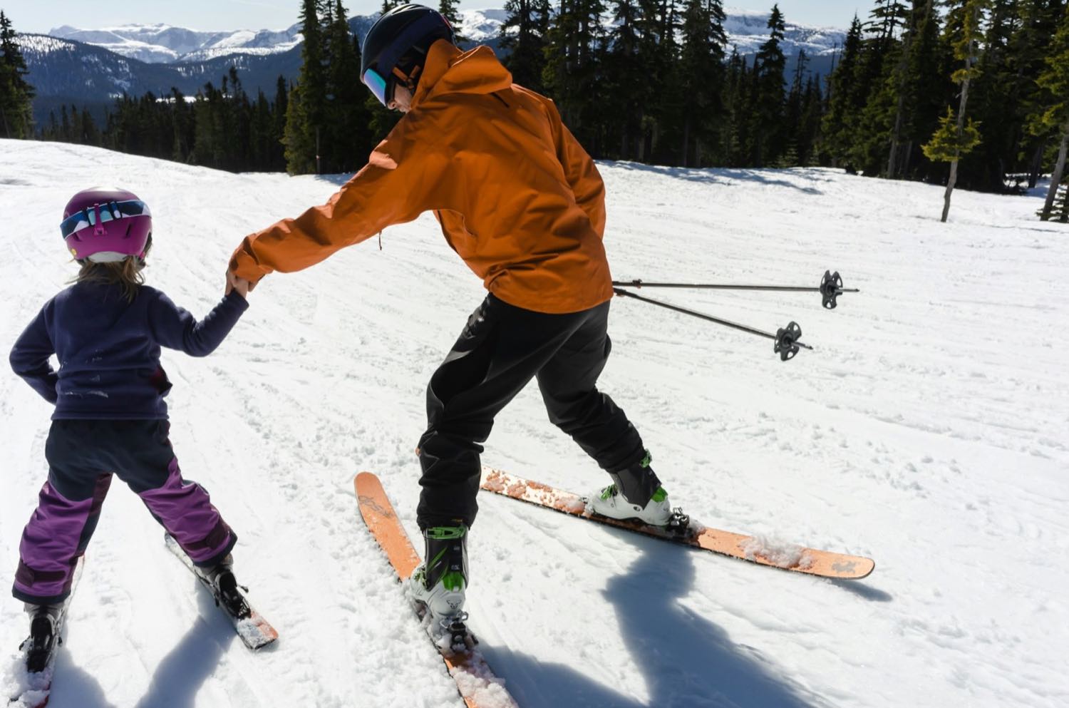 An adult in an orange jacket holding a small child’s hand as she learns to ski on a gentle slope